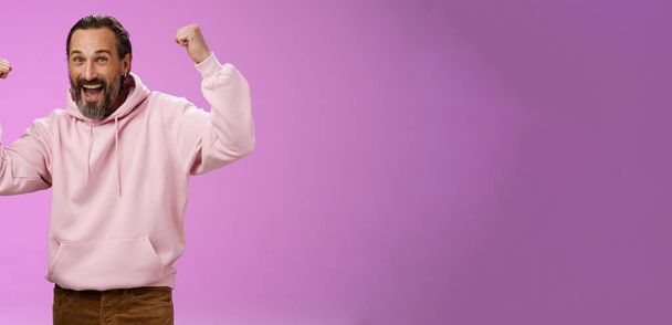Так, хто піклується про вік. Безтурботний задоволений щасливий старий бородатий у модному рожевому светрі піднімає кулаки радісно тріумфуючи весело, радіючи перемозі, відзначаючи успіх досягнення мети, позуючи фіолетовий фон
. - Фото, зображення