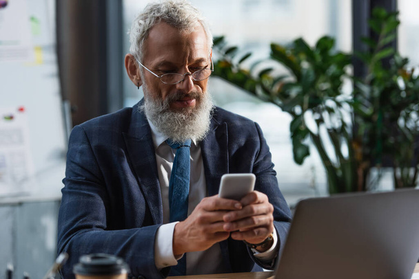Biznesmen w średnim wieku w okularach za pomocą smartfona w pobliżu laptopa i kawy w biurze  - Zdjęcie, obraz