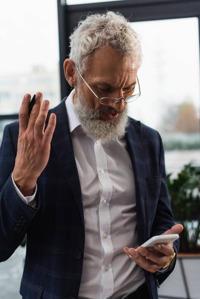 Uomo d'affari di mezza età in occhiali da vista utilizzando smartphone in ufficio  - Foto, immagini