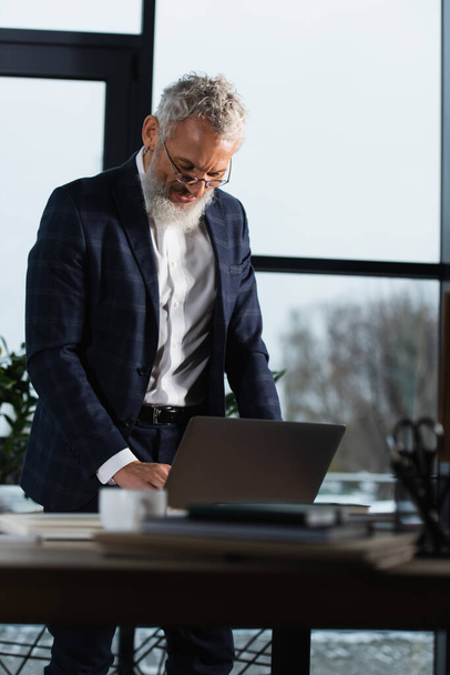 Mature manager in suit standing near laptop on table in office in evening  - Photo, image