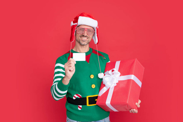Veselý vánoční dárek. muž držící debetní kartu pro nakupování. rád muž v elfím kostýmu a Santa klobouku s brýlemi. Vánoční chlapík s dárkovou krabicí na červeném pozadí. šťastný nový rok. - Fotografie, Obrázek
