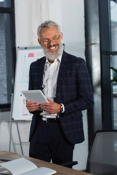 Cheerful grey haired businessman holding digital tablet near notebook on table in office  - Foto, Bild