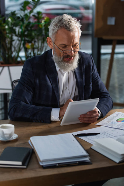 Homme d'affaires souriant aux cheveux gris utilisant une tablette numérique près des documents et du café au bureau  - Photo, image