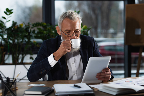 Бородатый бизнесмен в костюме пьет кофе и пользуется цифровой планшетом рядом с документами в офисе  - Фото, изображение