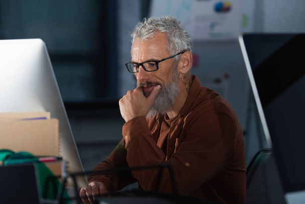 Focused businessman in eyeglasses using computer in office in evening  - Zdjęcie, obraz