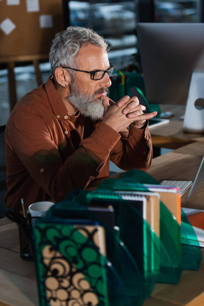 Focused mature businessman looking at computer monitor near notebooks in office in evening  - 写真・画像