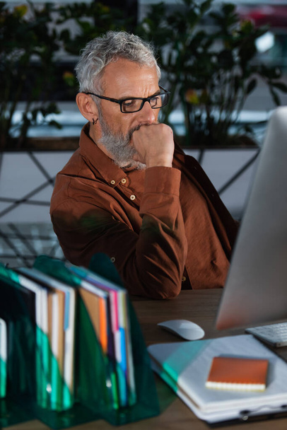 Geconcentreerde zakenman van middelbare leeftijd op zoek naar computer monitor bijna wazig papierwerk in het kantoor  - Foto, afbeelding