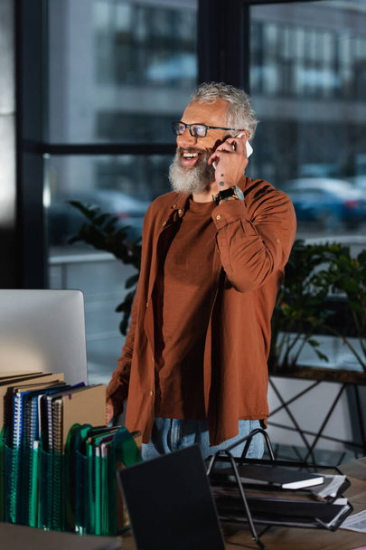 Cheerful grey haired manager talking on cellphone in office in evening  - Photo, image