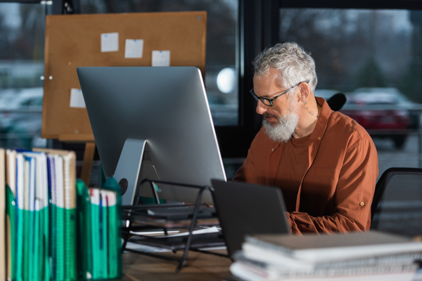 Empresário maduro em óculos trabalhando perto de computadores e documentos borrados no escritório  - Foto, Imagem