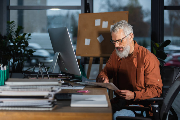 hombre de negocios barbudo en gafas que trabajan con documentos cerca de portátiles y monitor de computadora en la oficina por la noche - Foto, Imagen