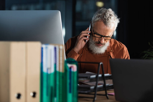hombre de negocios de pelo gris en gafas que hablan en el teléfono inteligente cerca de las computadoras y carpetas borrosas en la oficina por la noche - Foto, Imagen