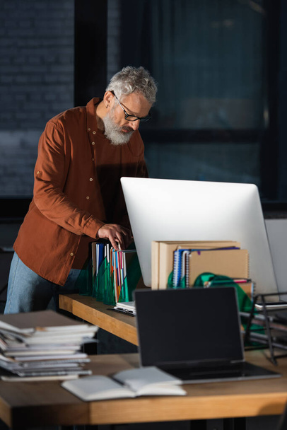 зрелый бизнесмен работает с бумагами возле компьютеров в темном офисе - Фото, изображение