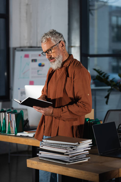 volwassen man met baard in bril staande met notebook op het werk in de buurt stapels documenten - Foto, afbeelding
