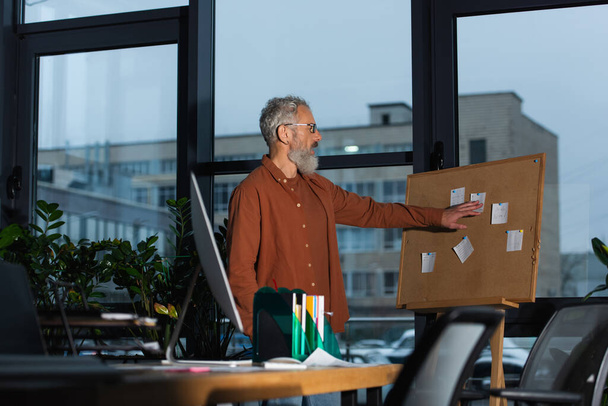 vista lateral do empresário de meia-idade olhando para notas de papel em cortiça enquanto trabalhava à noite no escritório - Foto, Imagem