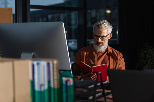 mature businessman in eyeglasses looking in notebook near computer monitor in dark office - Photo, image