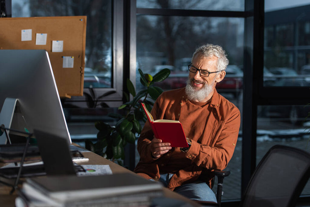 feliz hombre de negocios de mediana edad sentado con copybook cerca del monitor de la computadora en el lugar de trabajo en la oficina - Foto, imagen