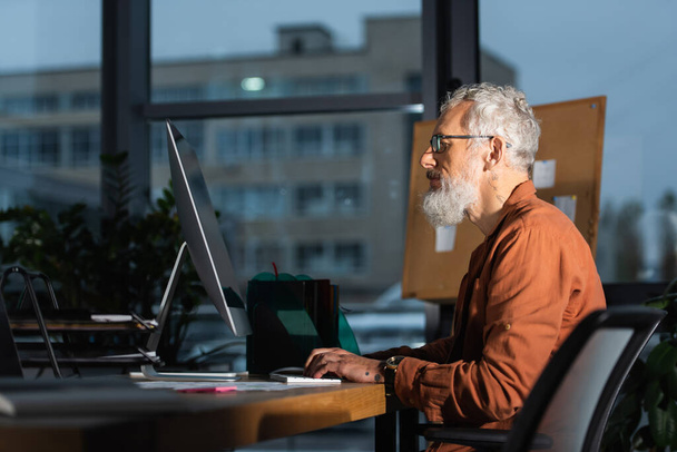 side view of bearded businessman in eyeglasses typing on laptop in office in evening - Фото, зображення