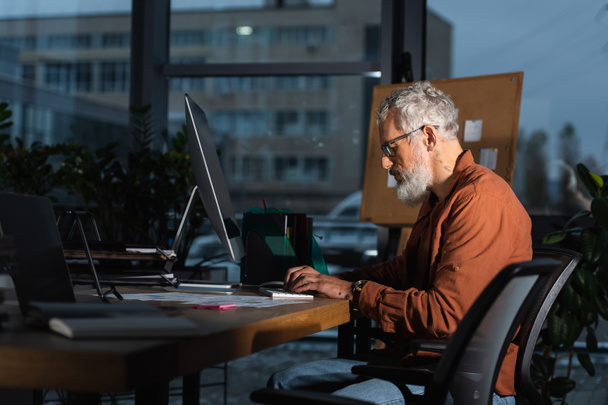 visão lateral de homem de negócios de meia idade barbudo trabalhando no computador no escritório à noite - Foto, Imagem
