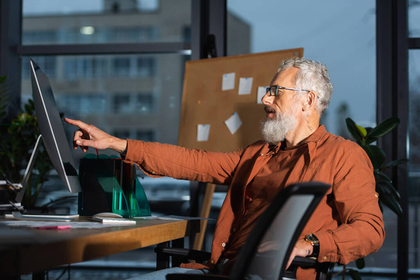 side view of bearded grey haired businessman pointing at computer monitor while sitting at workplace in office - Valokuva, kuva