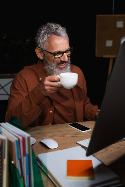 homme d'affaires joyeux avec tasse de café assis sur le lieu de travail près du smartphone et écran d'ordinateur flou - Photo, image