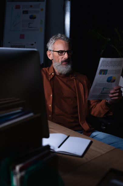 серьезный бородатый бизнесмен смотрит на бумагу с графиками, сидя рядом с пустым блокнотом ночью в офисе - Фото, изображение