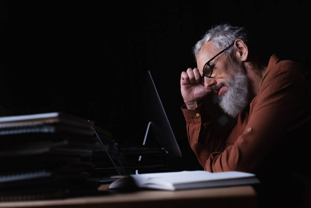 vista lateral del hombre de negocios de mediana edad cansado sentado cerca de computadoras y cuadernos borrosos en la oficina oscura - Foto, Imagen