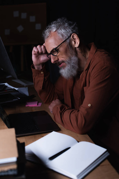 πλαϊνή άποψη του κουρασμένος και στοχαστικός επιχειρηματίας κάθεται στο φορητό υπολογιστή και κενό σημειωματάριο στο γραφείο τη νύχτα - Φωτογραφία, εικόνα