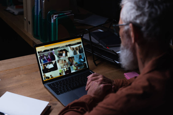 middelbare leeftijd zakenman werken op laptop in donker kantoor op wazig voorgrond - Foto, afbeelding