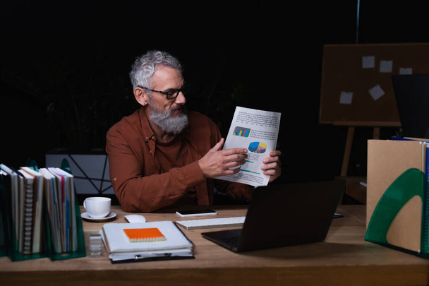 middle aged businessman holding paper with graphs during video chat on laptop in office at night - Photo, Image