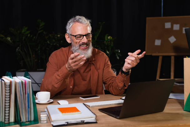 homme d'affaires barbu joyeux pointant vers un ordinateur portable tout en ayant appel vidéo au bureau la nuit - Photo, image
