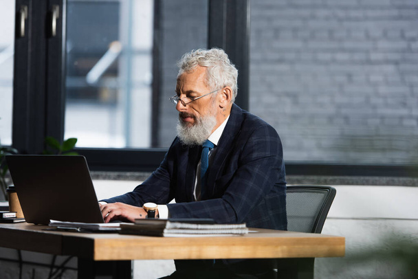 седой зрелый мужчина в формальной одежде работает на ноутбуке в офисе - Фото, изображение