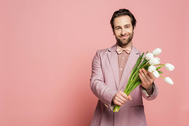 Бородатый ведущий мероприятия с белыми тюльпанами и глядя на камеру на розовом фоне  - Фото, изображение