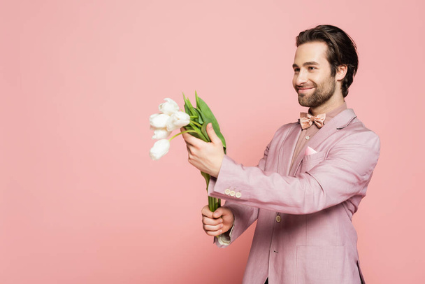 Positive host of event in jacket holding bouquet of tulips and looking away on pink background  - Photo, Image