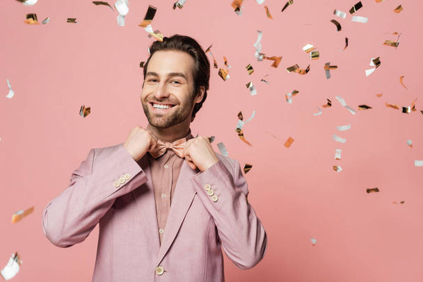 Smiling host of event in jacket adjusting bow tie under confetti on pink background  - Фото, изображение