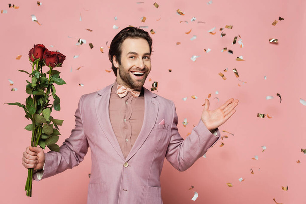 Ενθουσιασμένος οικοδεσπότης της εκδήλωσης κρατώντας τριαντάφυλλα και δείχνοντας με το χέρι κάτω από κομφετί σε ροζ φόντο  - Φωτογραφία, εικόνα