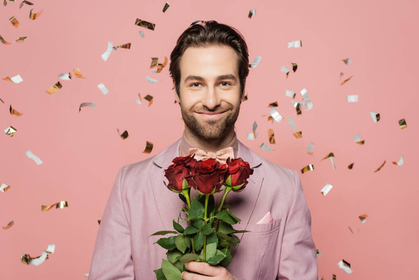 Portrait of positive host of event holding roses under falling confetti on pink background  - Φωτογραφία, εικόνα