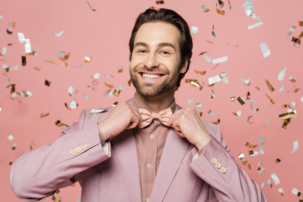 Smiling host of event adjusting bow tie near blurred confetti on pink background  - Fotografie, Obrázek