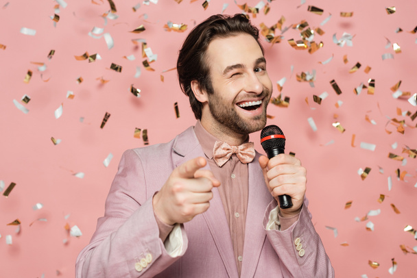 Positive host of event holding microphone and winking at camera under confetti on pink background  - Fotografie, Obrázek