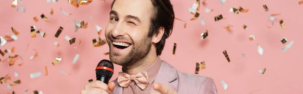 Stylish host of event holding microphone and winking at camera under confetti on pink background, banner  - Fotografie, Obrázek