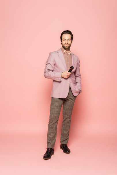 Full length of trendy host of event holding microphone and looking at camera on pink background  - Photo, Image