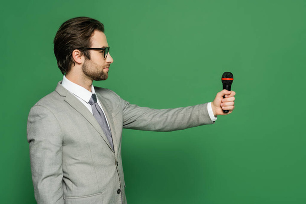 Oldalnézet hírolvasó öltönyben tartja mikrofon és félrenéz a zöld háttér  - Fotó, kép