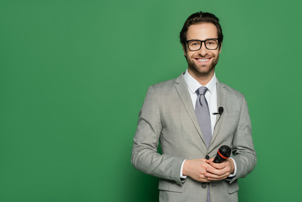 cheerful news anchor in suit and eyeglasses holding microphone on green background  - Photo, Image