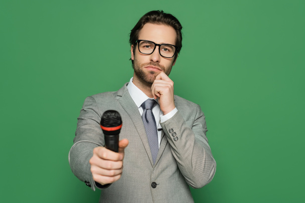 pensive άγκυρα ειδήσεων στο κοστούμι και γυαλιά που κρατούν μικρόφωνο απομονώνονται σε πράσινο  - Φωτογραφία, εικόνα