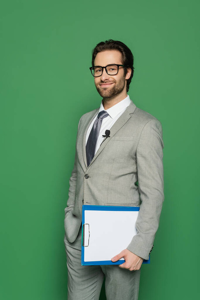 happy news anchor in suit and eyeglasses holding clipboard while standing with hand in pocket isolated on green  - Foto, Bild