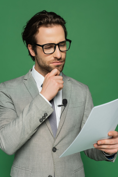 thoughtful news anchor in eyeglasses and suit looking at blank paper isolated on green - Photo, Image
