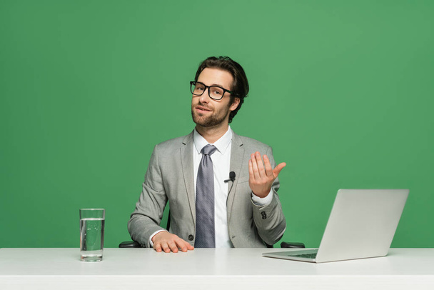 bebaarde nieuwslezer in bril en pak in de buurt van laptop geïsoleerd op groen - Foto, afbeelding