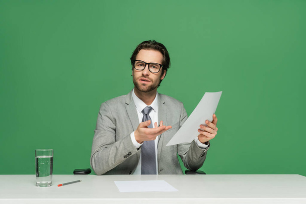 confuso presentador de noticias en anteojos y traje apuntando con la mano a papel en blanco aislado en verde - Foto, Imagen