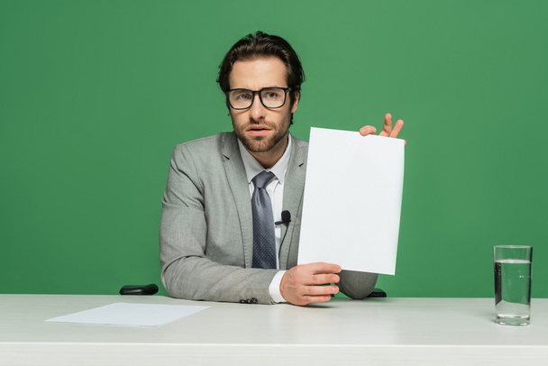 Бородатый ведущий новостей в очках и костюме, держащий чистую бумагу, изолированную на зеленом - Фото, изображение