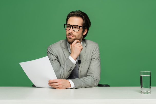 вдумчивый телеведущий в очках и костюмах держит чистую бумагу, изолированную на зеленом - Фото, изображение