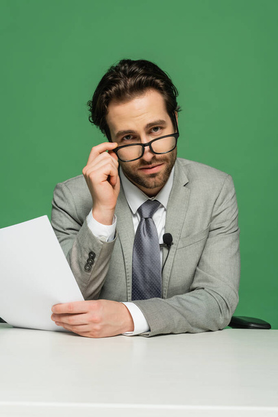 broadcaster in suit adjusting eyeglasses and holding blank paper isolated on green - Photo, Image
